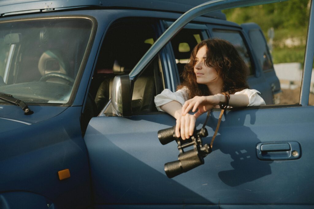 Woman with binoculars enjoying nature from SUV window in a serene outdoor setting.